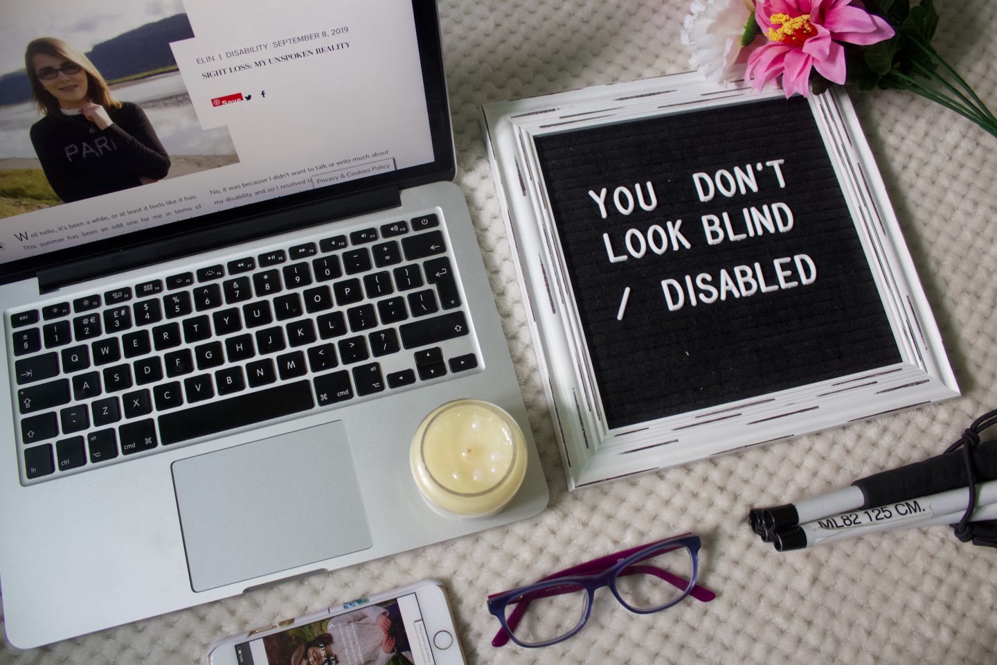A photo of one of my blog posts on a macbook screen, there's a small unlit candle at the edge of the keyboard and a felt letter board beside it that says "You don't look blind /disabled"