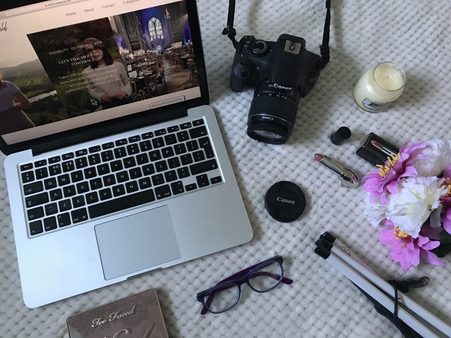 A photo of my blog's homepage on a macbook, there's a canon camera beside it, a pair of glasses, a white cane and some pink and white artificial flowers