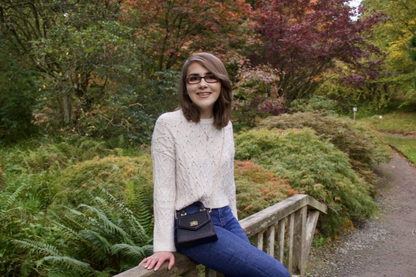 A photo of Elin sitting on a small wooden bridge, she is smiling at the camera and there are pretty trees and flowers in the background, all in pretty autumnal colours