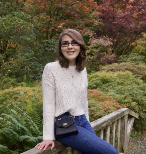 A photo of Elin sitting on a small wooden bridge, she is smiling at the camera and there are pretty trees and flowers in the background, all in pretty autumnal colours
