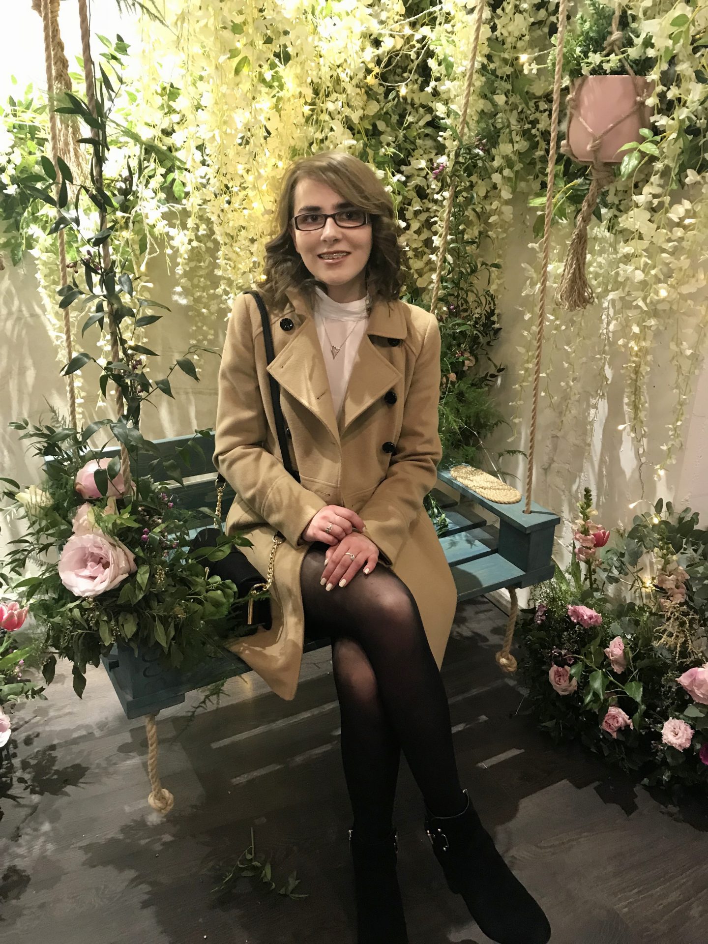 A photo of Elin sitting on a seat swing in the Duck and Dry salon, there are flowers behind and around her and she is wearing a camel coat over an ivory pleated blouse and black denim skirt. Her short hair is curled and she is smiling at the camera