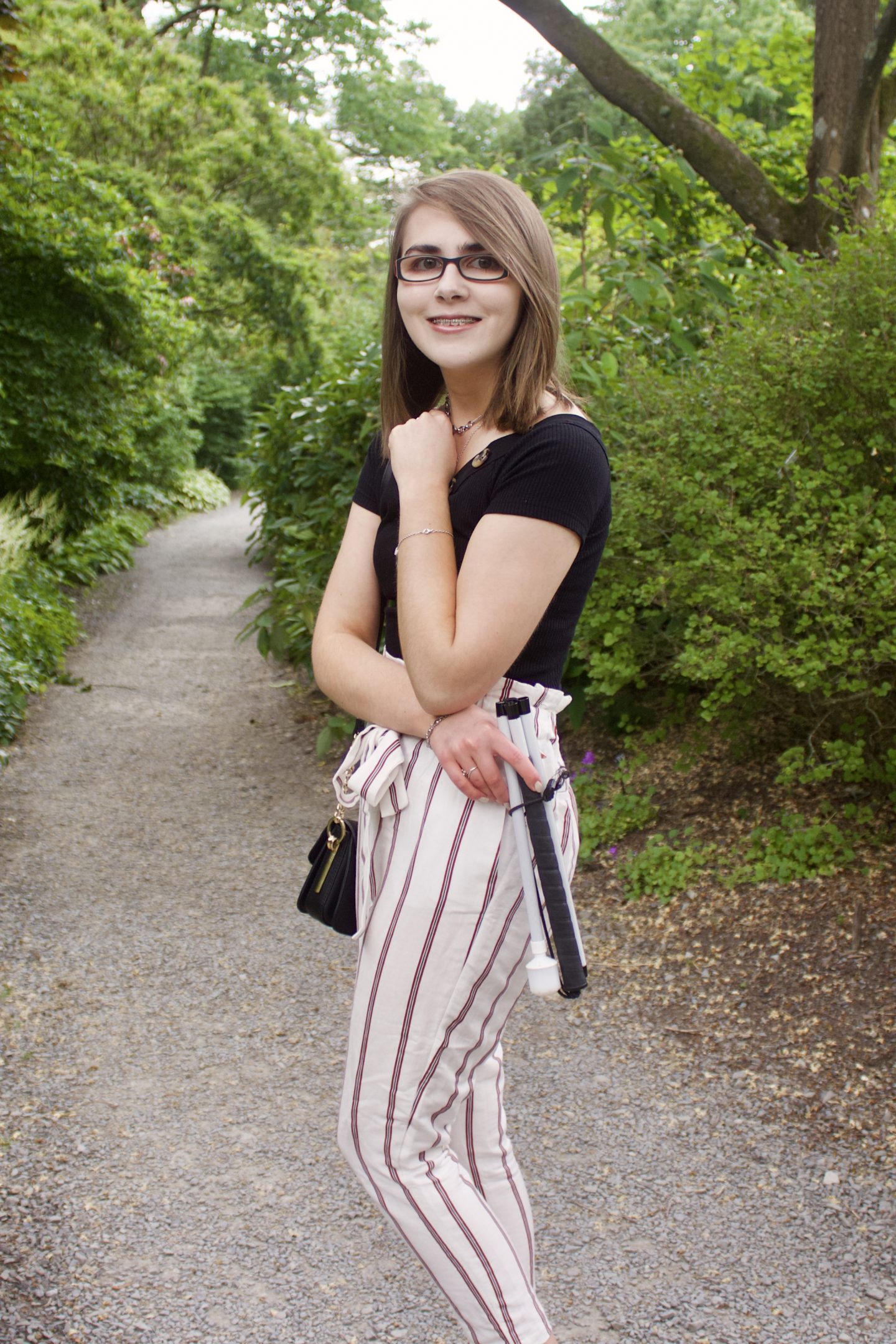 A photo of Elin standing on a path which leads through trees and flowers. She is wearing skinny cream trousers with red stripes, a black bardot top with buttons on the neckline and a black handbag