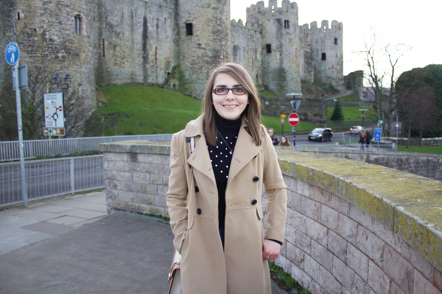 A photo of Elin standing in front of a castle, she is wearing a double breasted camel coat, a black high neck top which has silver embellishment on the front and dark blue skinny jeans