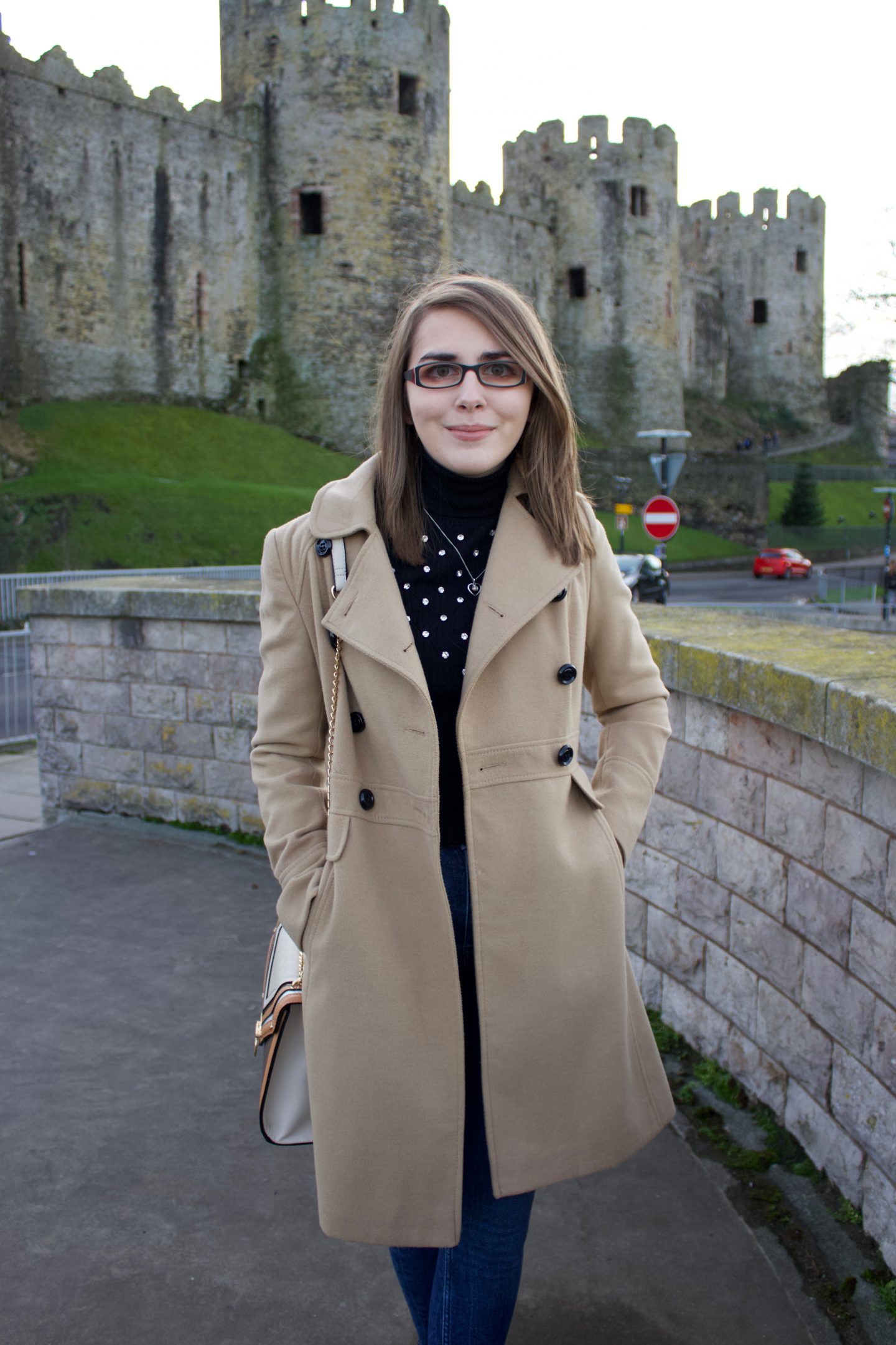 A photo of Elin standing in front of a castle, she is wearing a double breasted camel coat, a black high neck top which has silver embellishment on the front and dark blue skinny jeans