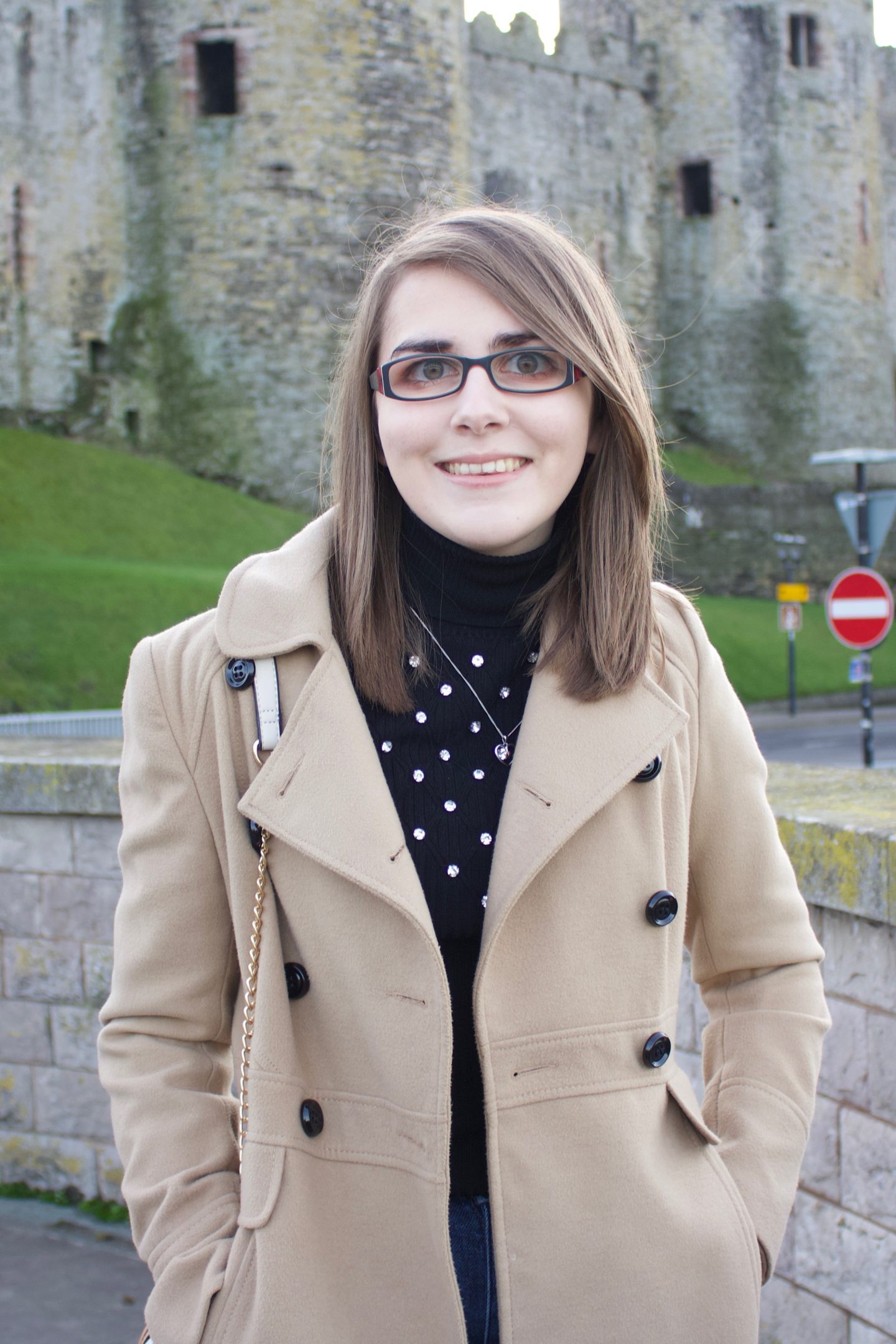 A photo of Elin standing in front of a castle, her hands are in her coat pockets and she is smiling at the camera