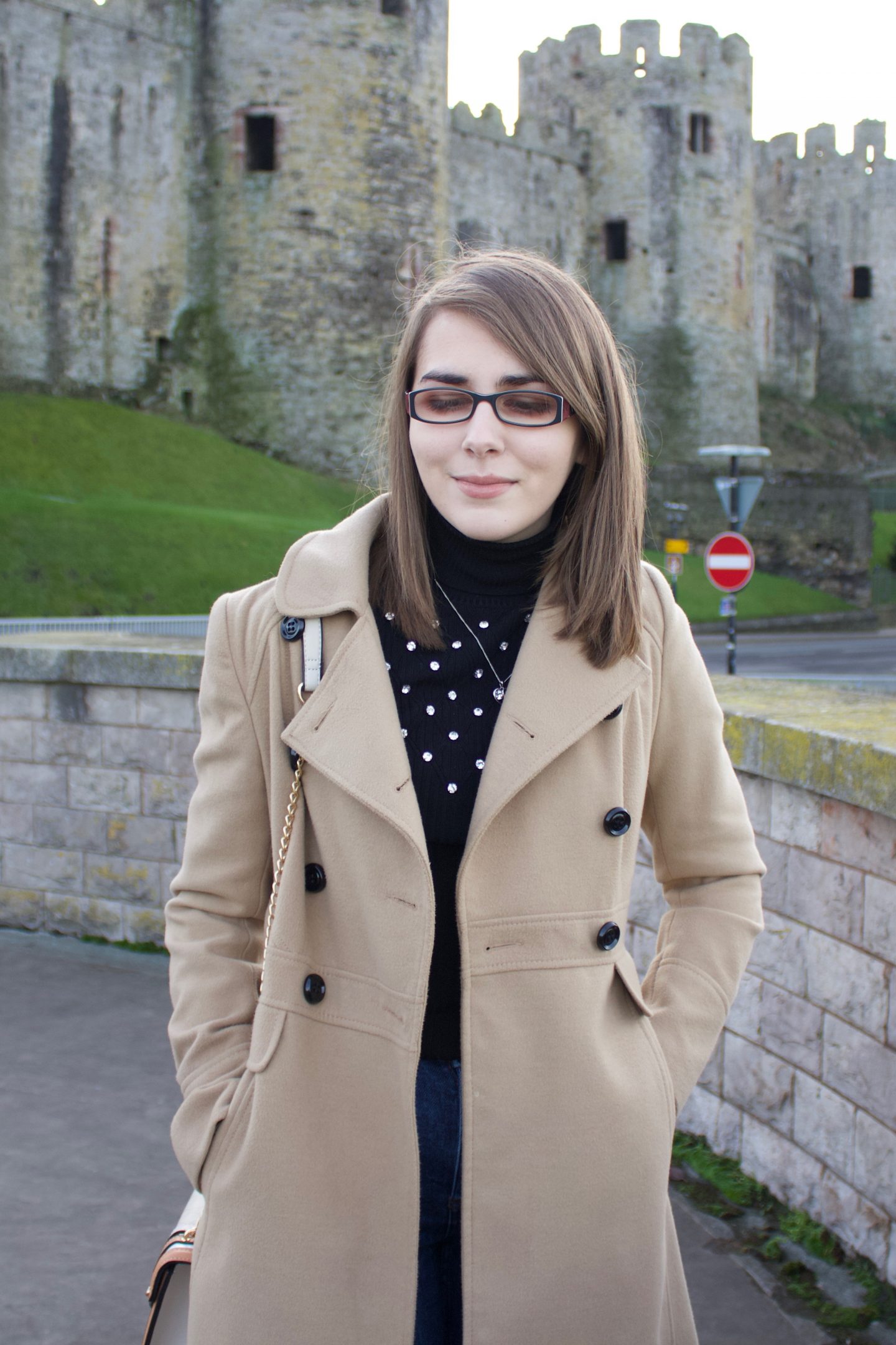 A photo of Elin standing in front of a castle, she is wearing a black high neck top, a camel coat and dark blue skinny jeans and she is looking down