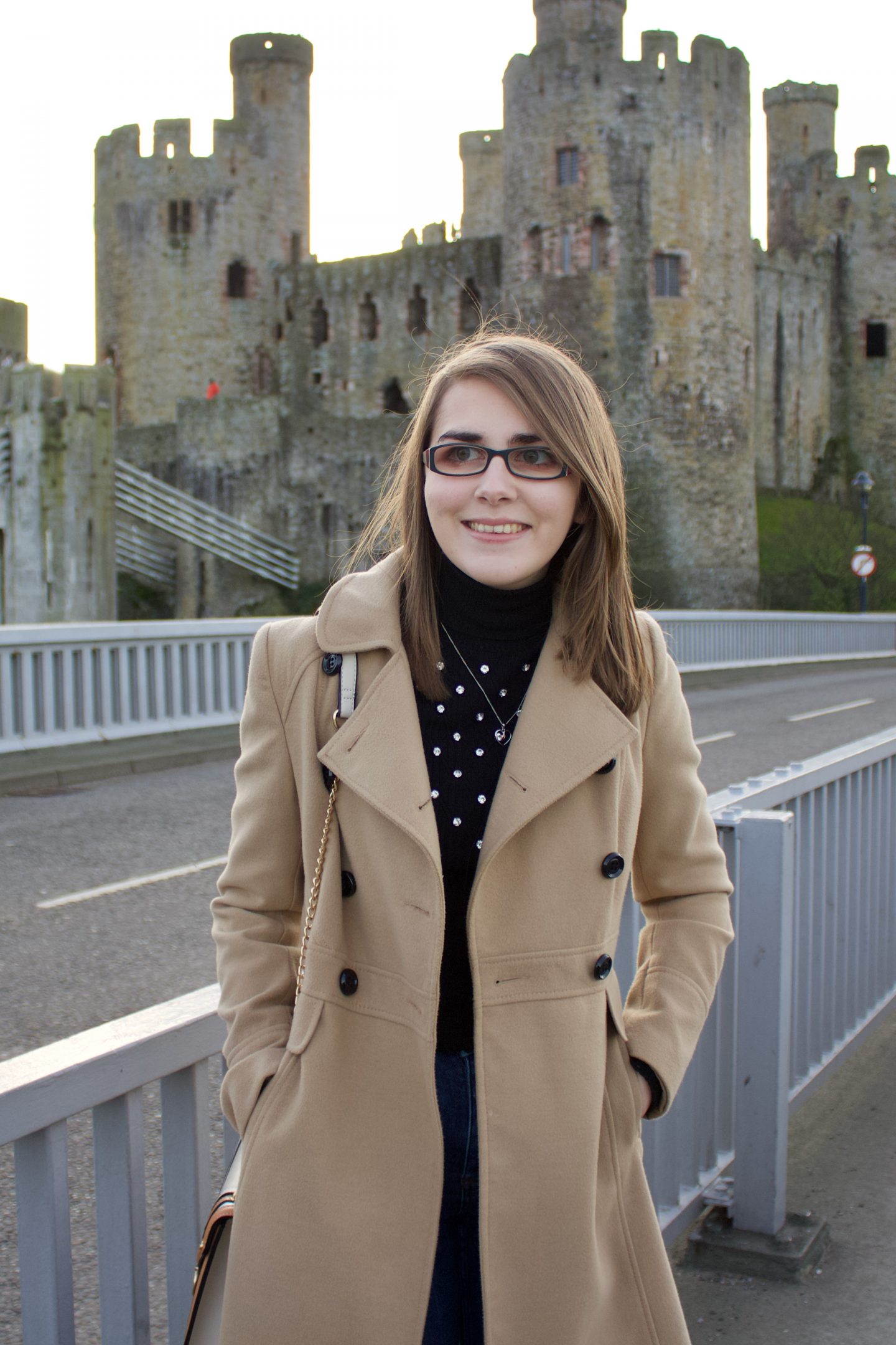 A photo of Elin standing on a bridge in front of a caslte, she is smiling and looking away from the camera