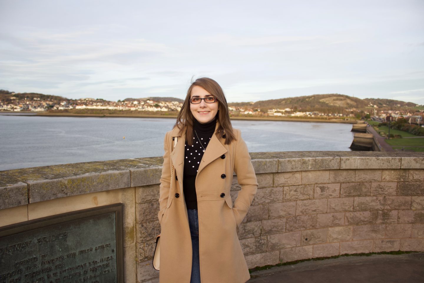 A photo of Elin standing in front of a view of a river with houses in the background. She is wearing a longline camel coat, a black high neck top which has embellishment on the front and dark blue skinny jeans. She's smiling at the camera