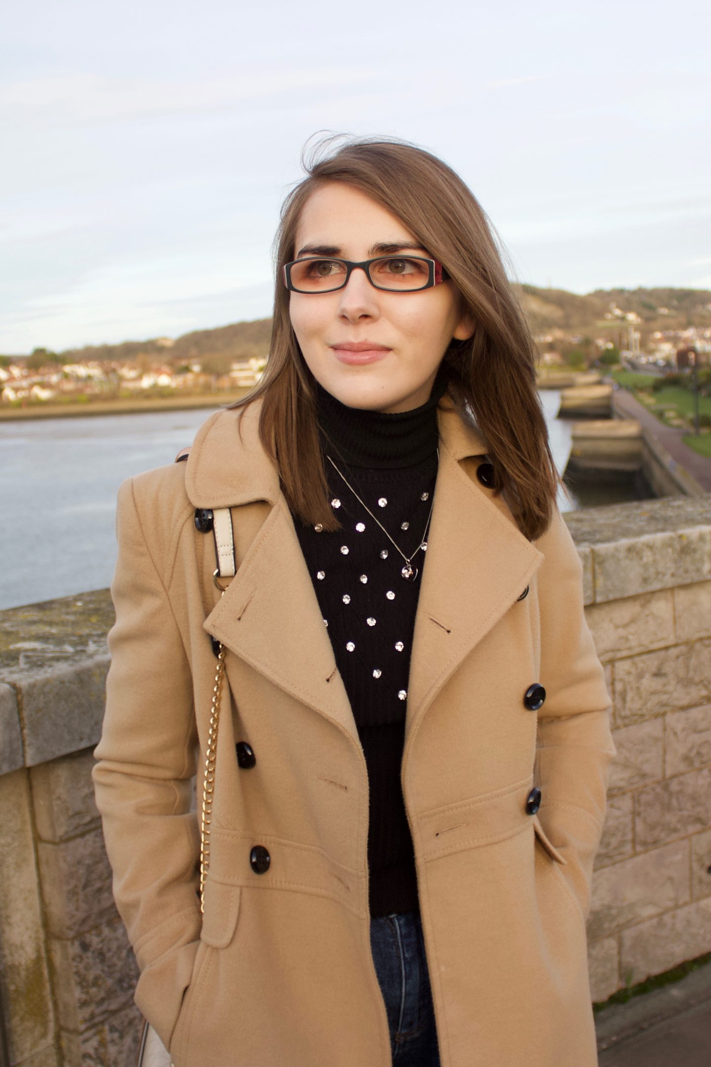 A photo of Elin standing in front of a river with houses on the other side of it, she is looking away from the camera. The sun is setting which creates golden hour lighting in the photo