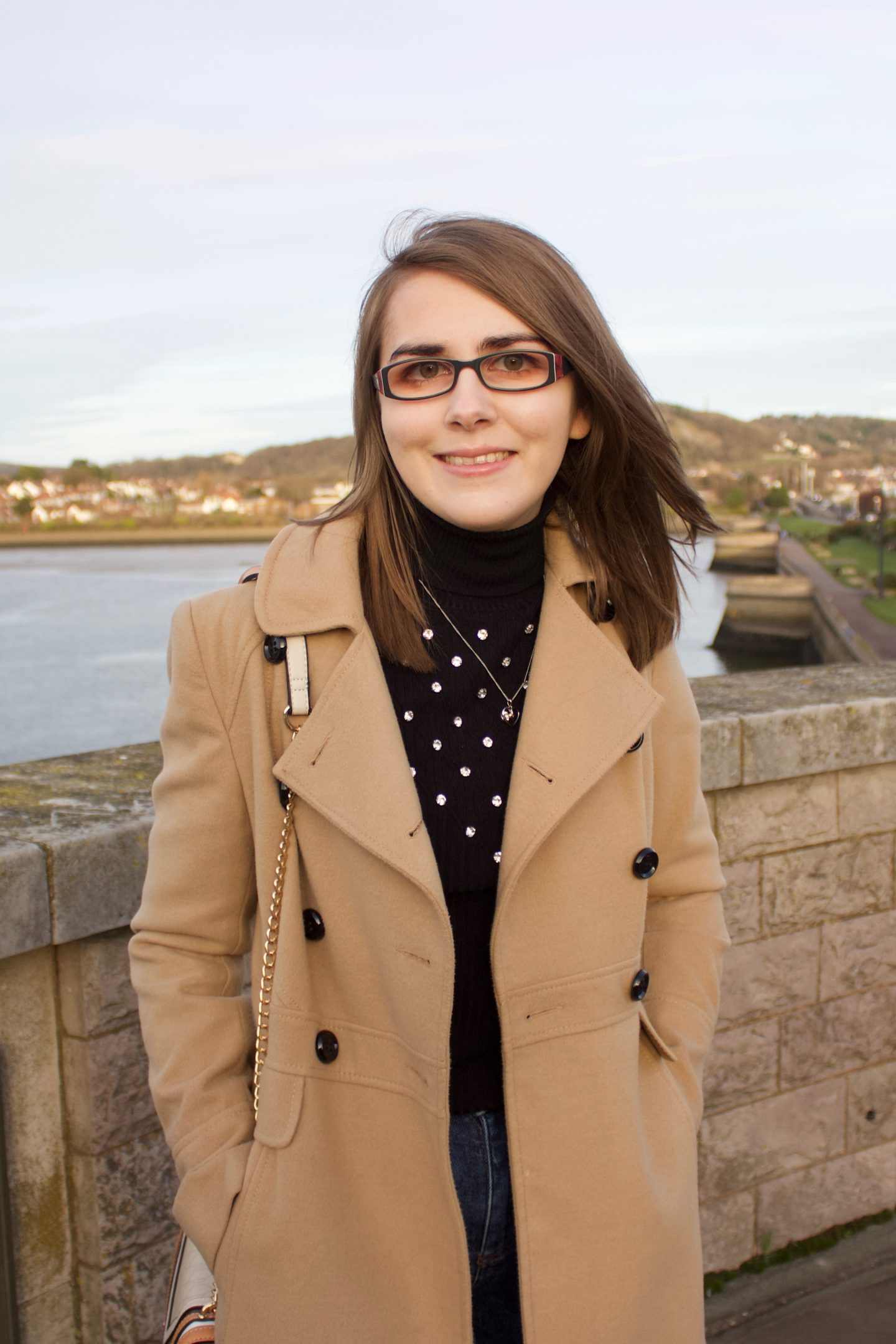 A photo of Elin standing in front of a river with houses in the background, she is smiling at the cameara and her hair is slightly windswept