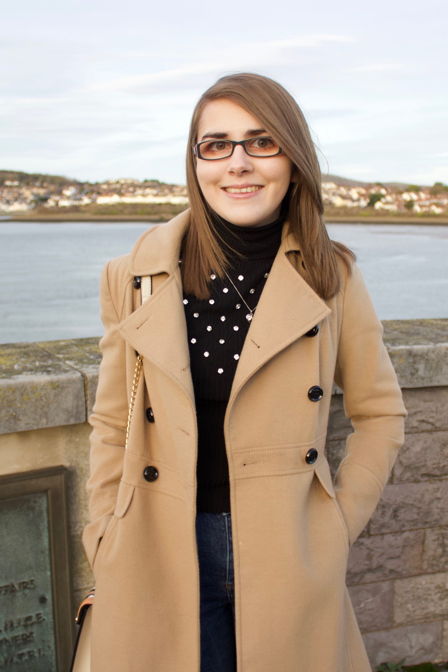 A photo of Elin standing in front of a river with houses on the other side of it, she is smiling at the camera. The sun is setting which creates golden hour lighting in the photo