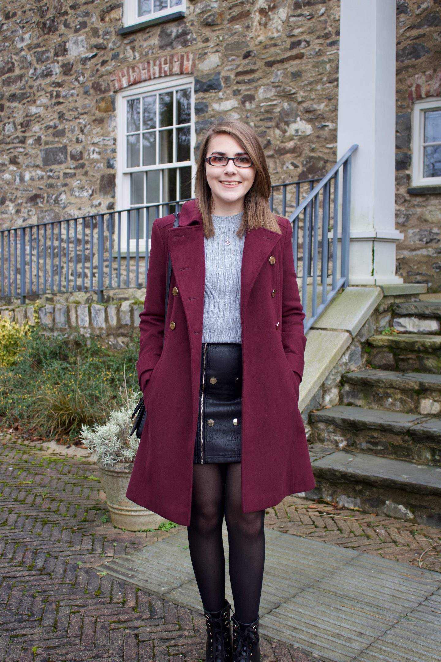 A photo of Elin wearing a faux black leather skirt which has a zip and button detailing on the front, she's paired it with a cropped grey knitted jumper, a burgundy coat, tights and ankle boots