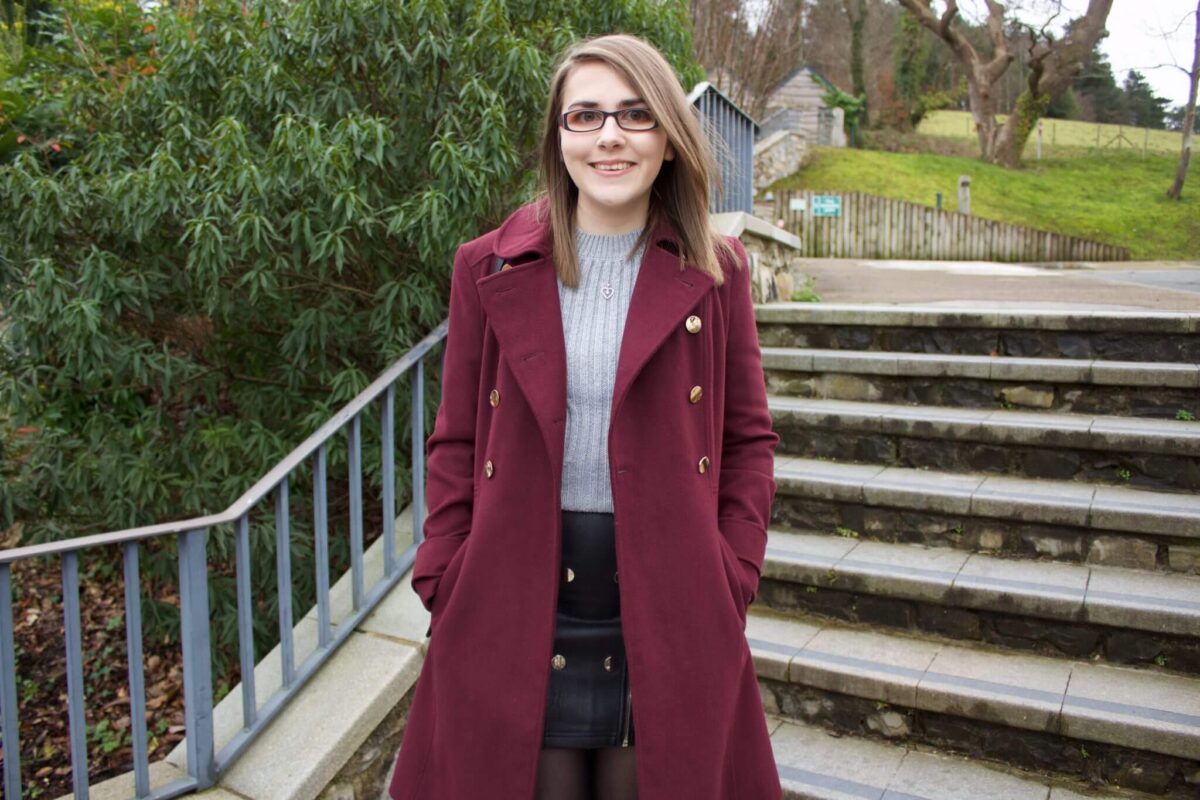 A photo of Elin standing on some stone steps, she is wearing a grey knitted jumper and black faux leather mini skirt which has buttons and zip detailing on the front. She is smiling at the camera