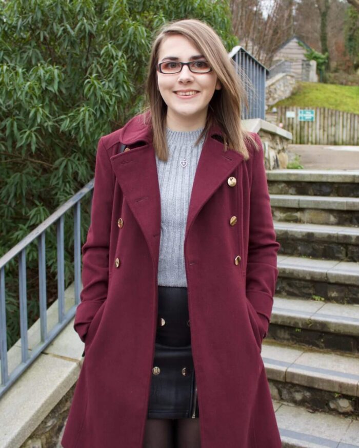 A photo of Elin standing on some stone steps, she is wearing a grey knitted jumper and black faux leather mini skirt which has buttons and zip detailing on the front. She is smiling at the camera