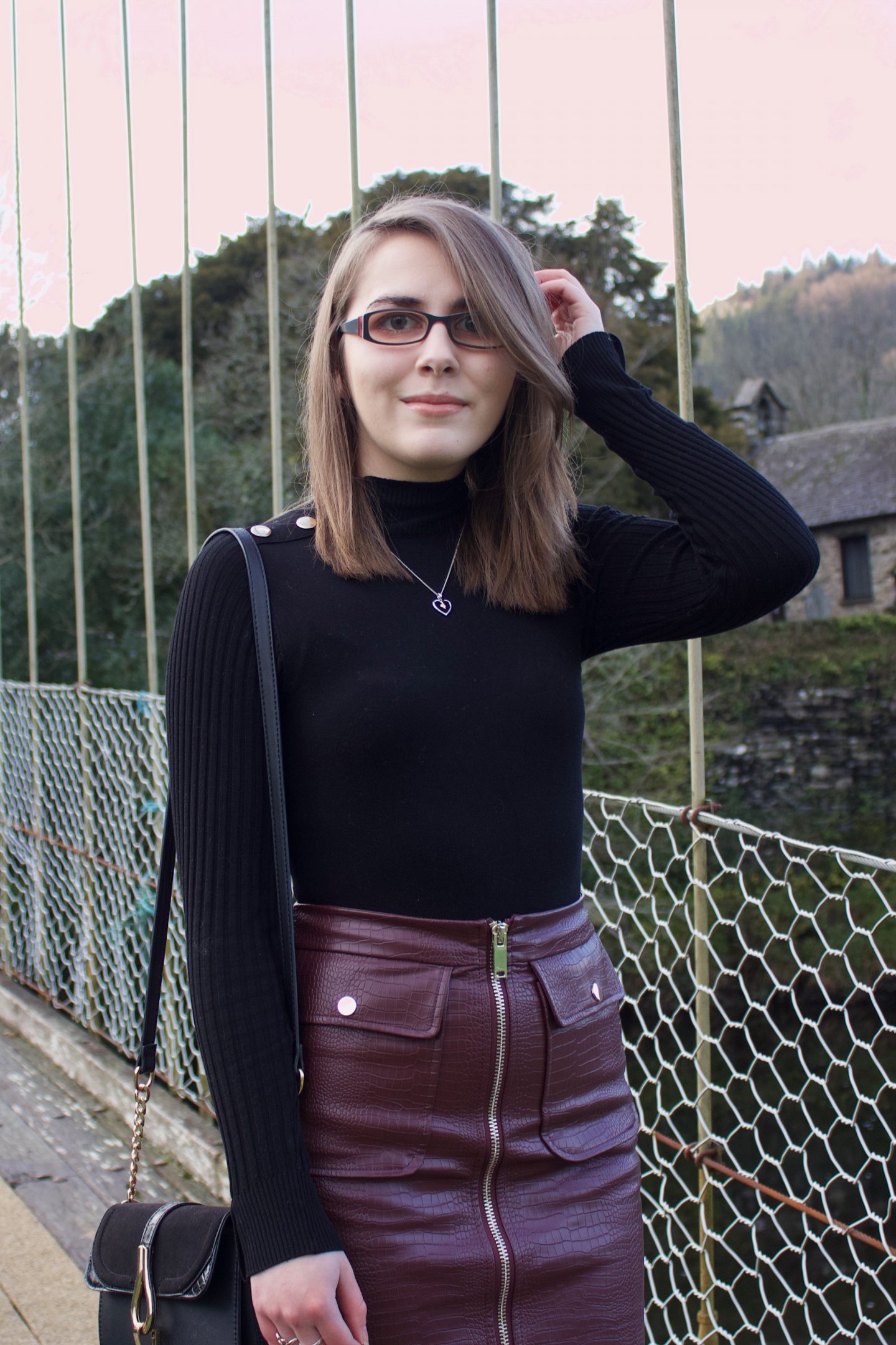 A photo of Elin stnding on a bridge, her left hand is raised to her hair and she's looking at the camera