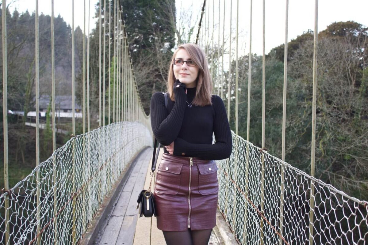 A photo of Elin standing on a suspension bridge with trees in the background. She wears a black high neck top which has gold buttons on the right shoulder and on the cuffs. It's paired with a burgundy faux leather mini skirt which has a zip in the middle and pockets either side