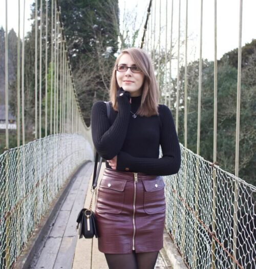 A photo of Elin standing on a suspension bridge with trees in the background. She wears a black high neck top which has gold buttons on the right shoulder and on the cuffs. It's paired with a burgundy faux leather mini skirt which has a zip in the middle and pockets either side