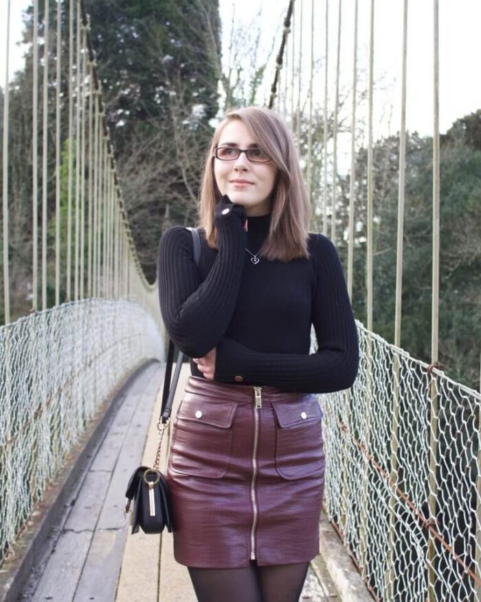 A photo of Elin standing on a suspension bridge with trees in the background. She wears a black high neck top which has gold buttons on the right shoulder and on the cuffs. It's paired with a burgundy faux leather mini skirt which has a zip in the middle and pockets either side