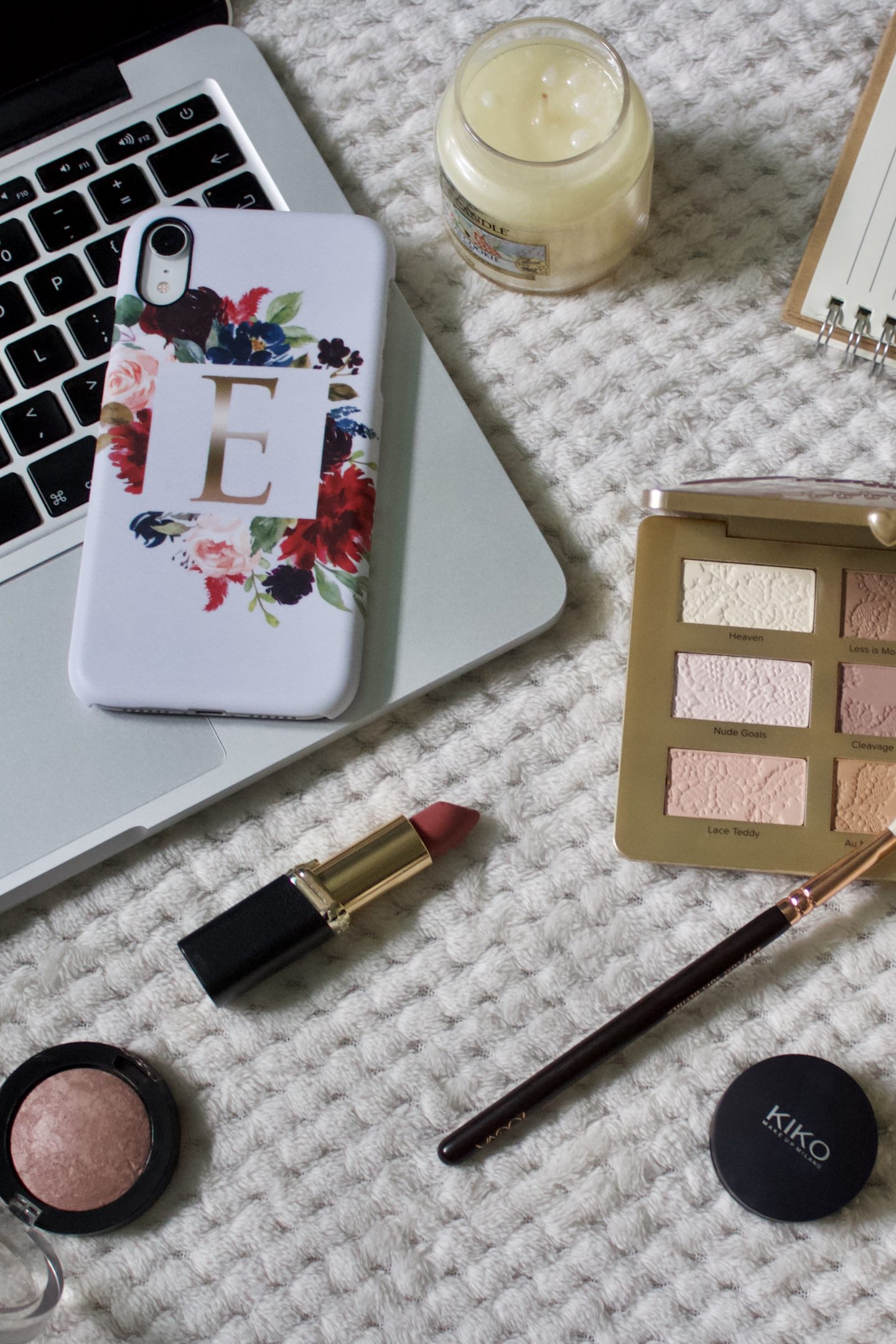 A photo of a white phone case which has flowers surrounding a printed initial, the case is placed on the edge of a macbook keyboard and there are various makeup products surrounding it including a lipstick and an eyeshadow palette