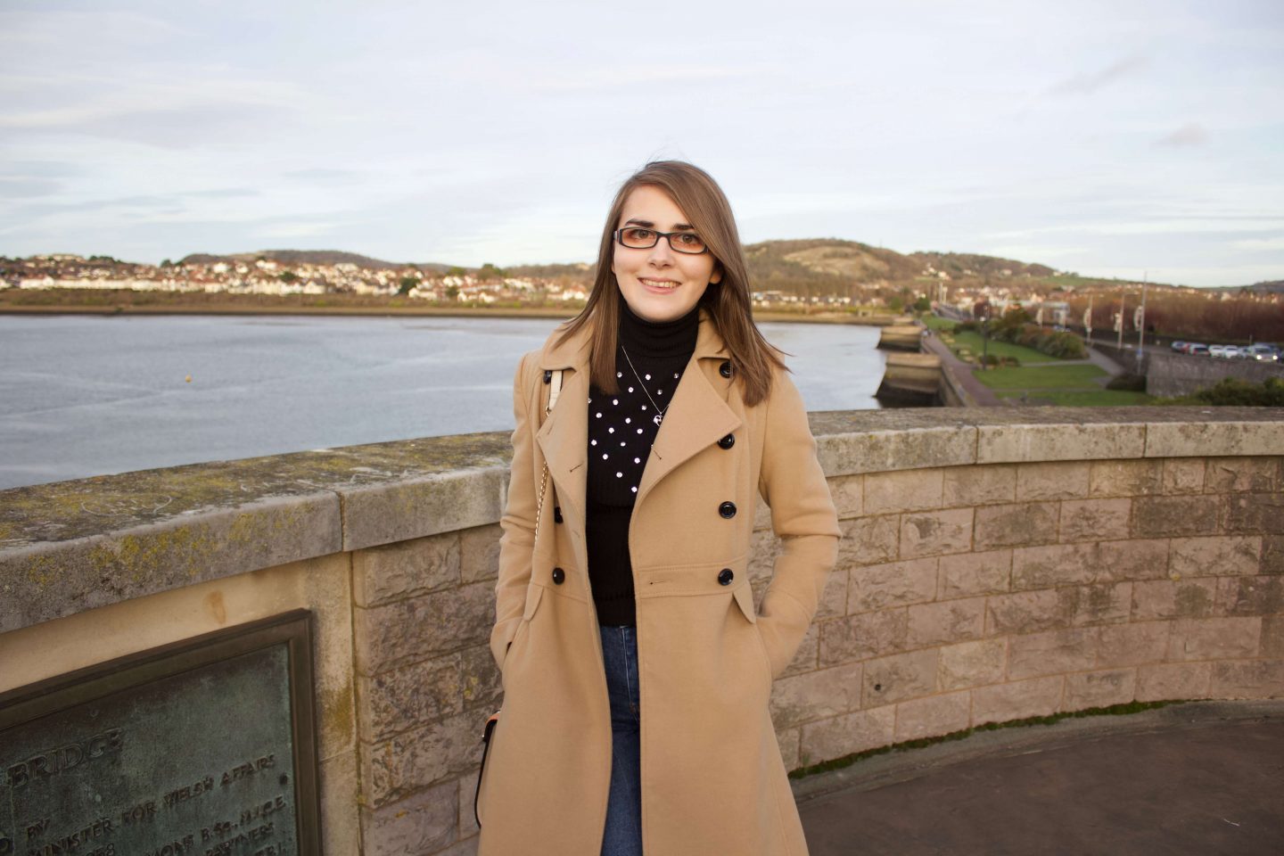 A photo of Elin standing in front of a view of a river with houses in the background. She is wearing a longline camel coat, a black high neck top which has embellishment on the front and dark blue skinny jeans. She's smiling at the camera. The sun is setting which creates golden hour lighting in the photo