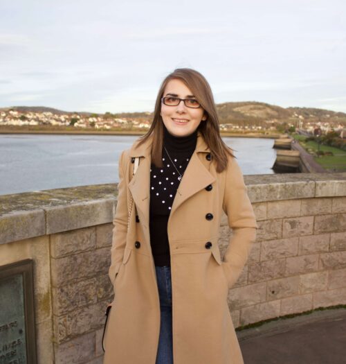 A photo of Elin standing in front of a view of a river with houses in the background. She is wearing a longline camel coat, a black high neck top which has embellishment on the front and dark blue skinny jeans. She's smiling at the camera. The sun is setting which creates golden hour lighting in the photo