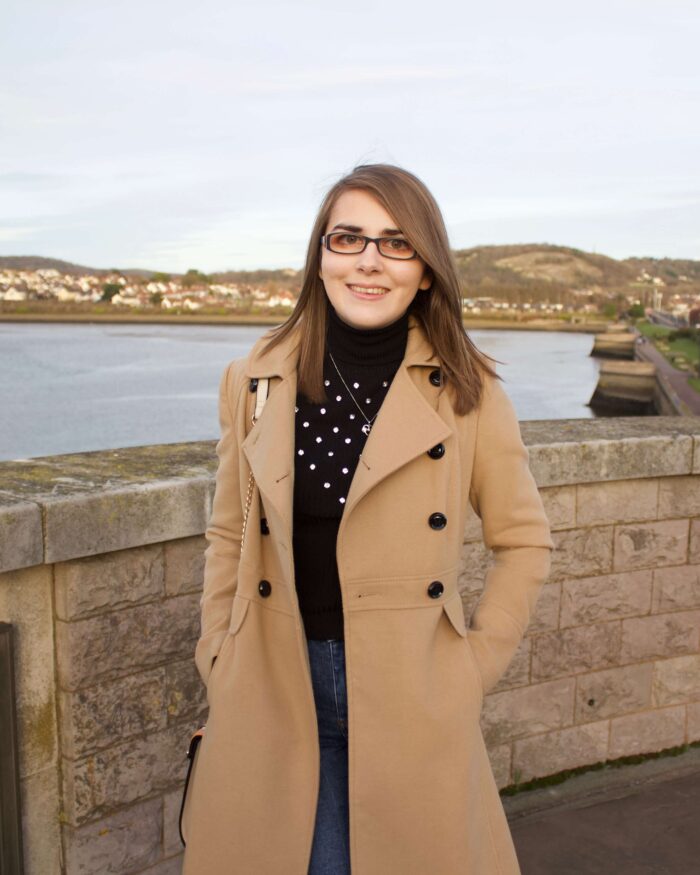 A photo of Elin standing in front of a view of a river with houses in the background. She is wearing a longline camel coat, a black high neck top which has embellishment on the front and dark blue skinny jeans. She's smiling at the camera. The sun is setting which creates golden hour lighting in the photo