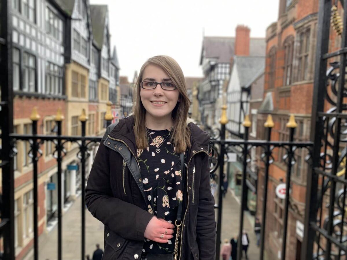 A photo of Elin wearing a black floral dres with a black coat, she's standing in front of pretty black railings overlooking a street of shops and pretty tudor style buildings