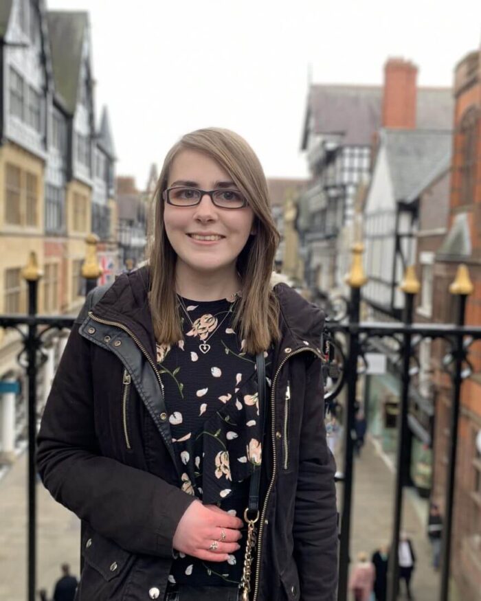 A photo of Elin wearing a black floral dres with a black coat, she's standing in front of pretty black railings overlooking a street of shops and pretty tudor style buildings