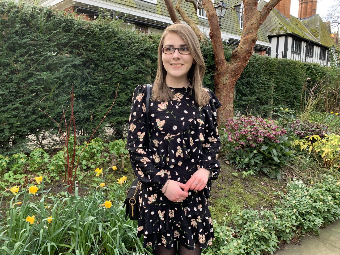 A photo of Elin standing in front of flowers and trees. A white and black tudor style house can be seen in the background. She's wearing a black floral dress which has ruffle detailing and she's looking away from the camera and smiling