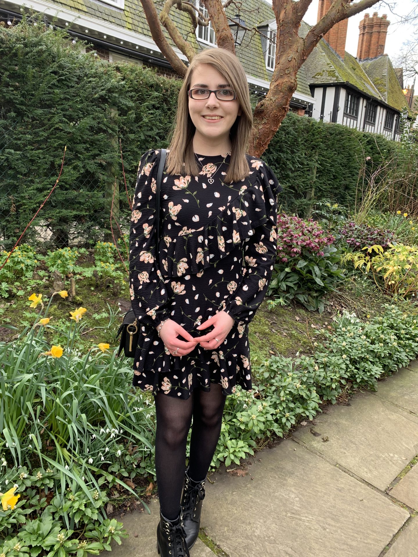 A photo of Elin wearing a black floral dress with ruffle detailing on the front. She's paired it with tights and black lace up ankle boots