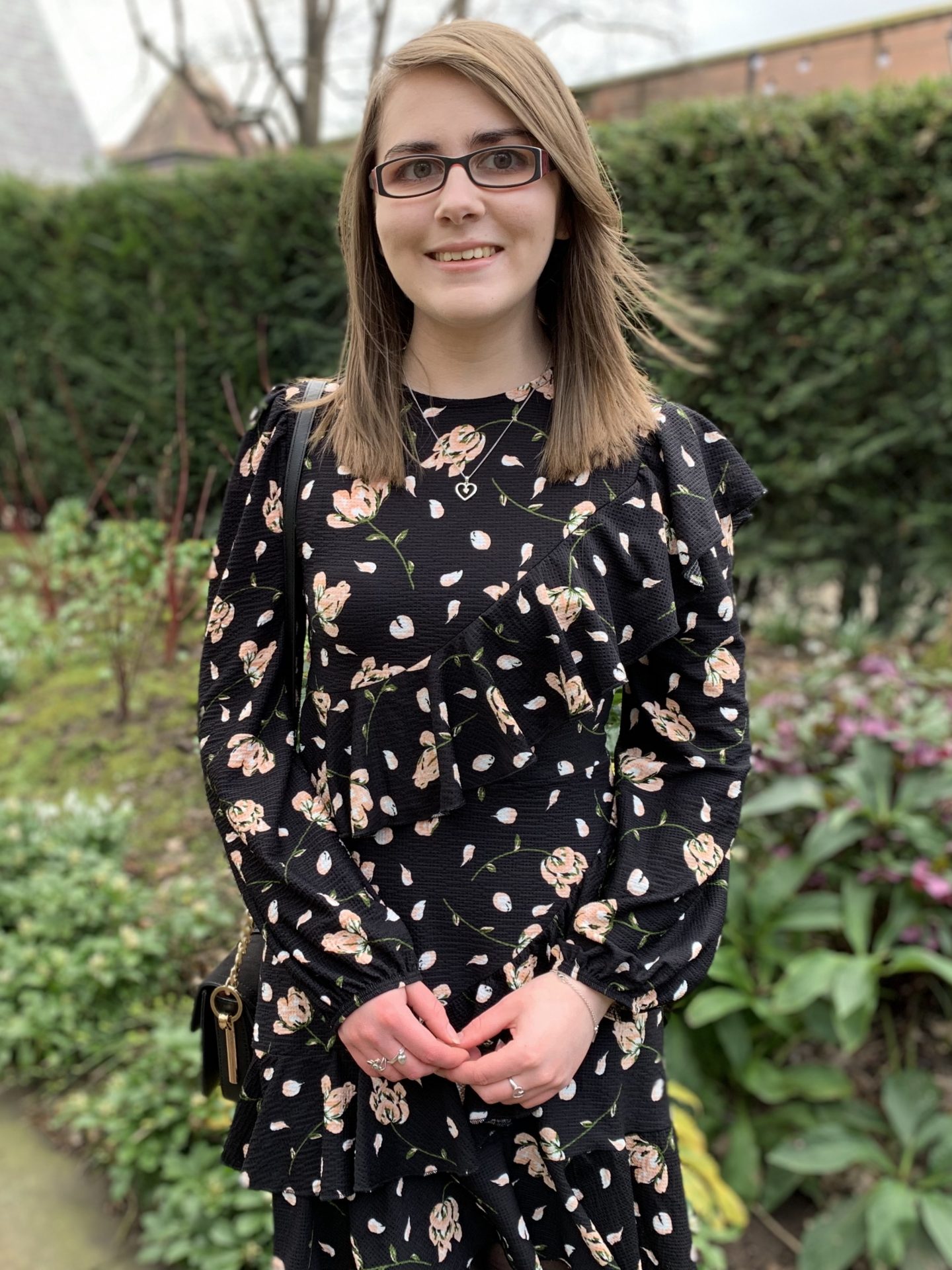 A photo of Elin standing in front of flowers with some buildings peeking out behind the hedge behind her. She's wearing a black floral dress and a silver heart shaped necklace. She's smiling at the camera