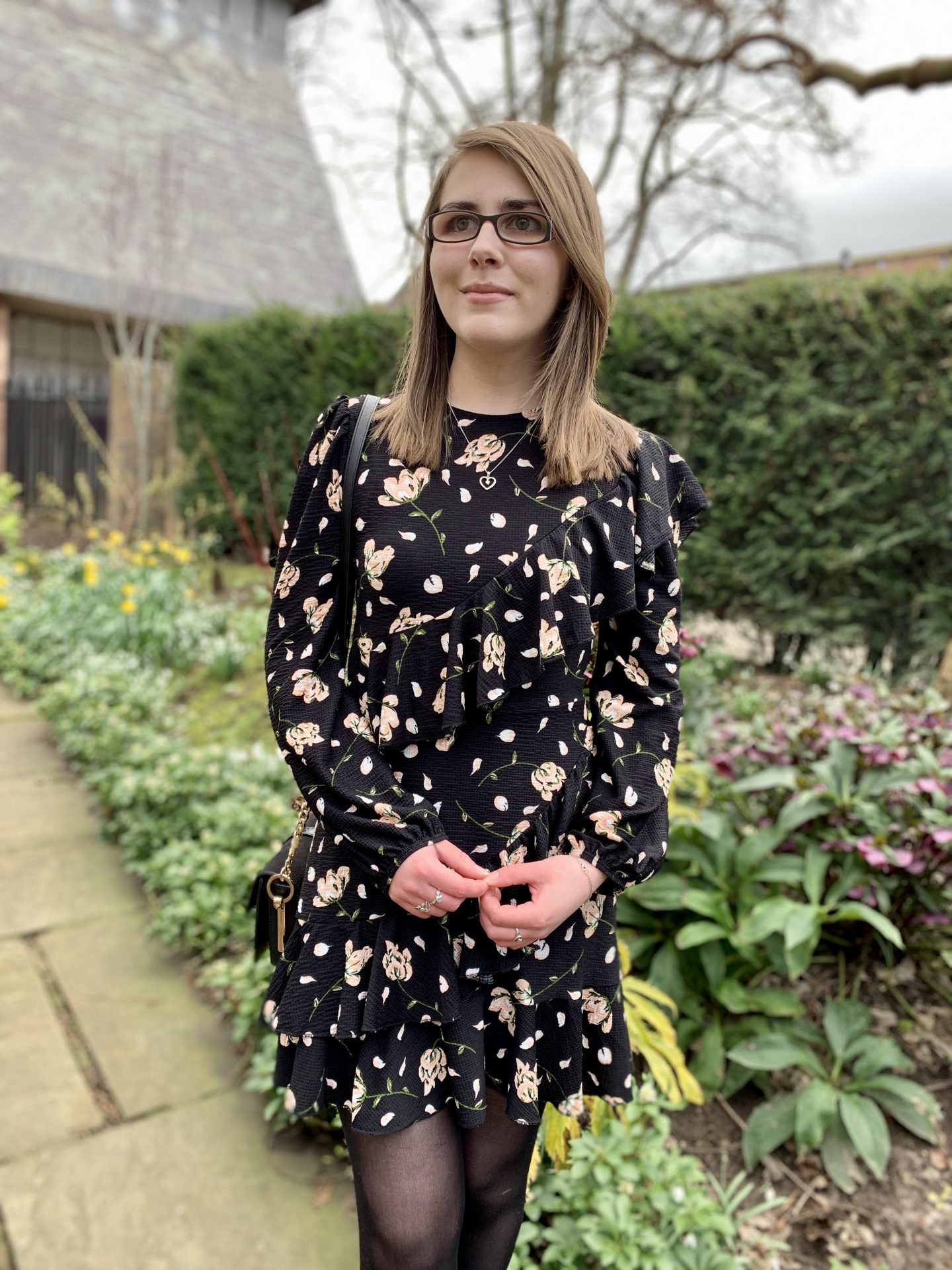 A photo of Elin looking away from the camera. She's standing in front of flowers with some buildings peeking out behind the hedge behind her. She's wearing a black floral dress and a silver heart shaped necklace.