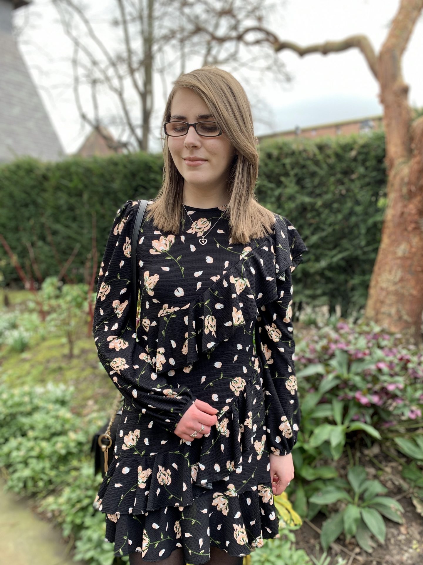 A photo of Elin standing in front of flowers with some buildings peeking out behind the hedge behind her. She's wearing a black floral dress and a silver heart shaped necklace. She's looking down
