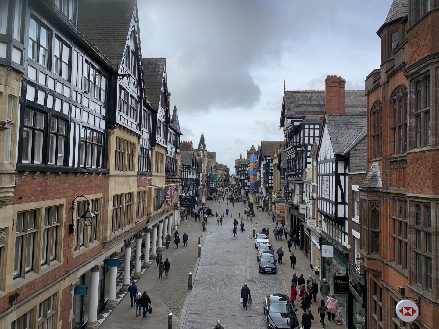 A photo of a street of shops in Chester from a high viewpoit. The street is strewn with people and there are pretty tudor style buildings along the street