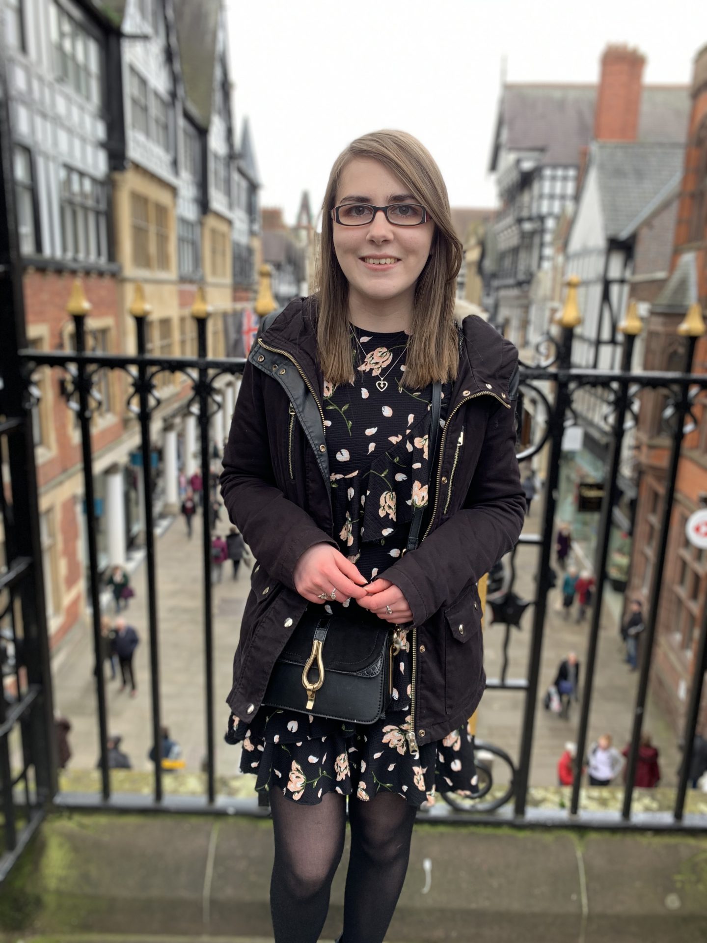 A photo of Elin standing in front of pretty black railings overlooking a street of shops and pretty buildings. She's wearing a black floral dress with a black coat and a little black bag