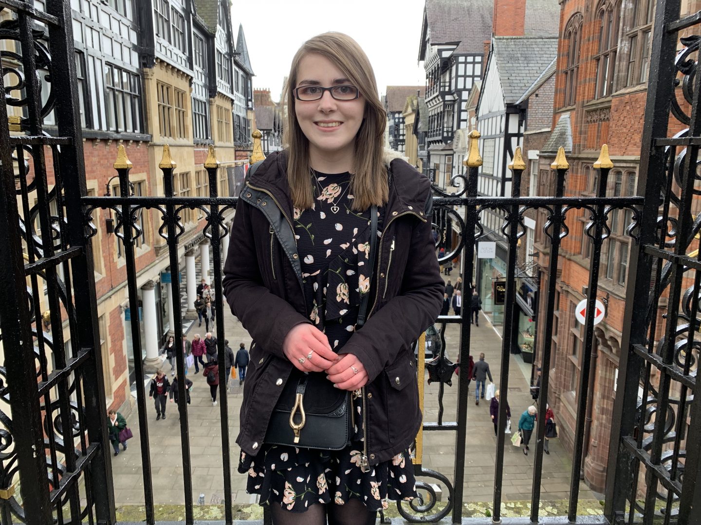 A photo of Elin standing in front of pretty black railings overlooking a street of shops and pretty buildings. She's wearing a black floral dress with a black coat and a little black bag