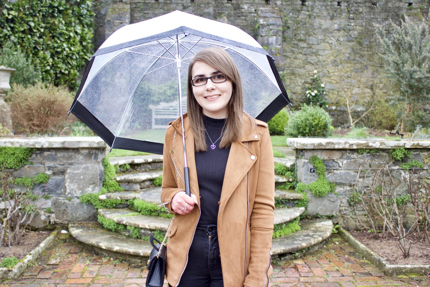 A photo of Elin standing on some stone steps in front of a pretty stone wall with flowers in front ot it. She's smiling at the camera