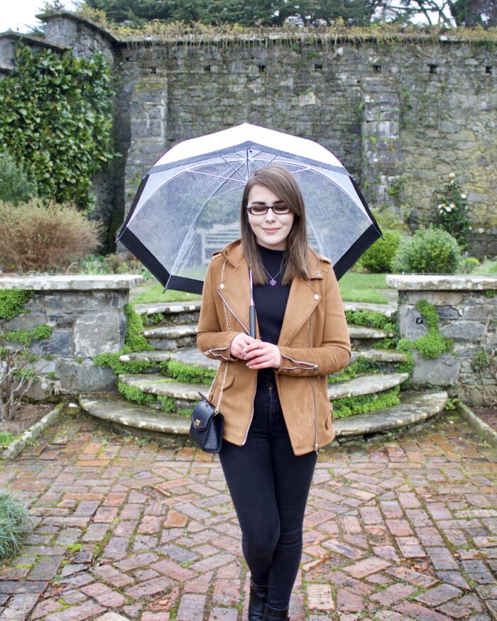 A photo of Elin standng in front of some stone steps with flowers around them. She's wearing a black high neck top and skinny jeans with a brown faux suede jacket