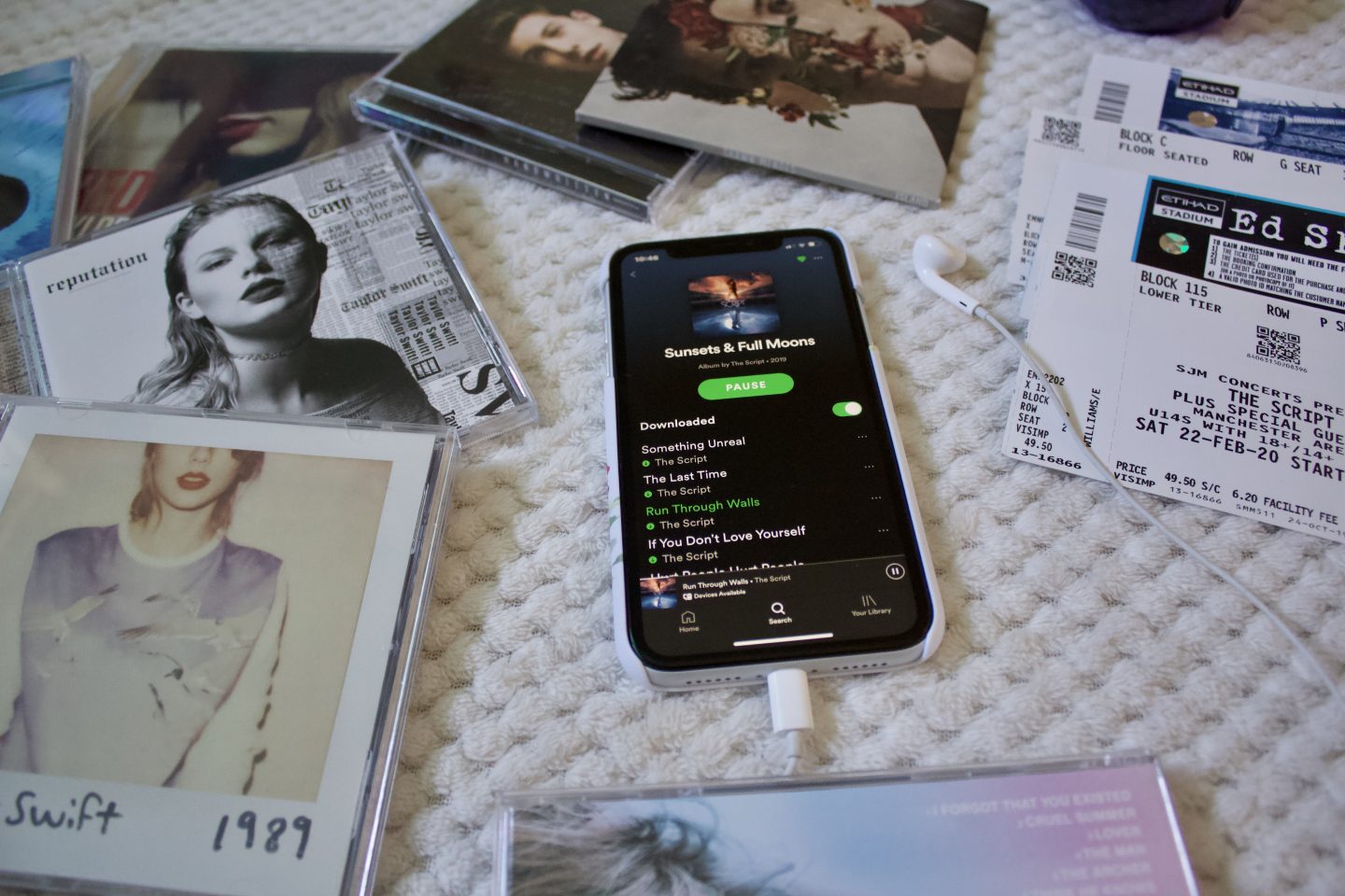 A photo of The Scripts Sunsets and full moons album playlist on an iPhone, its surrounded by concert tickets, earphones and a few albums including a few by Taylor Swift and Shawn Mendes