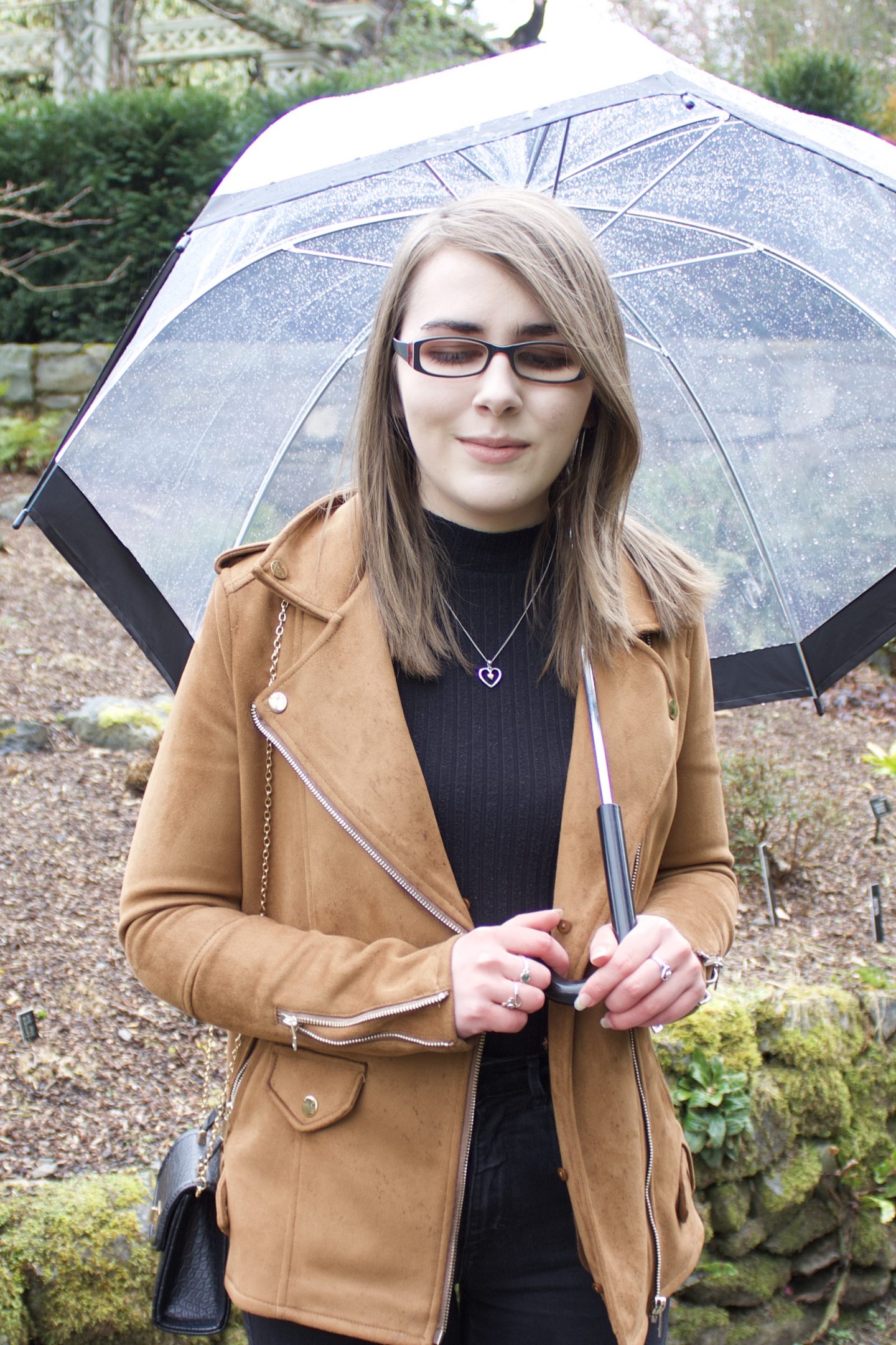 A photo of Elin wearing a brown faux suede jacket and black high neck top, she's holding a clear umbrella and looking down