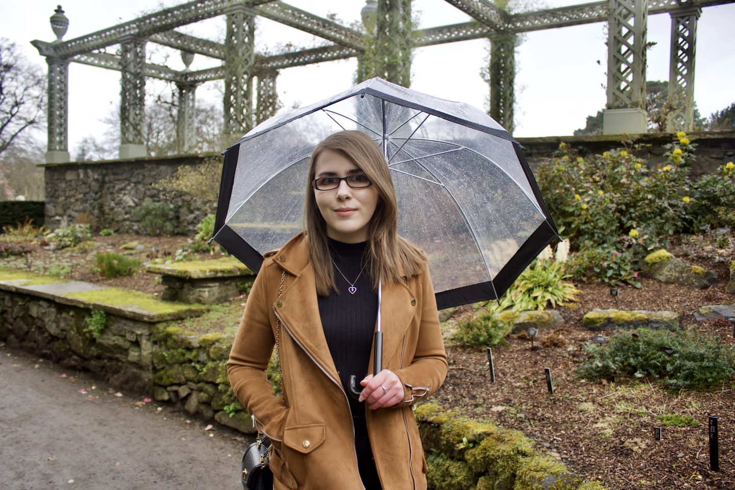 A photo of Elin standing in front of flower beds, she wears a black high neck top and a brown faux suede jacket. She's also holding a clear umbrella which has a black rim