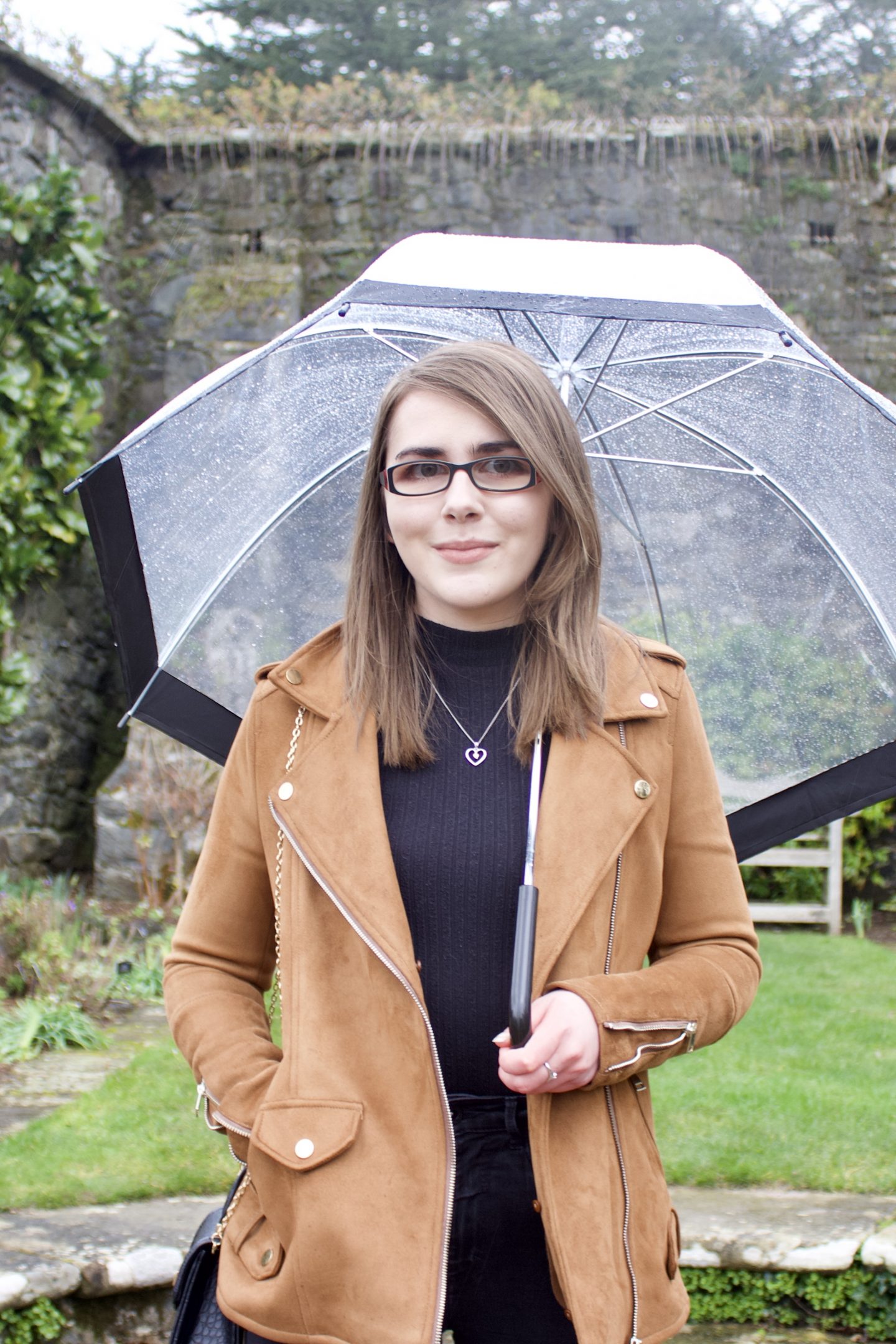 A photo of Elin standing on some stone steps in front of a pretty stone wall and a white metal bench. She wears a black high neck top, black skinny jeans and a brown suede jacket