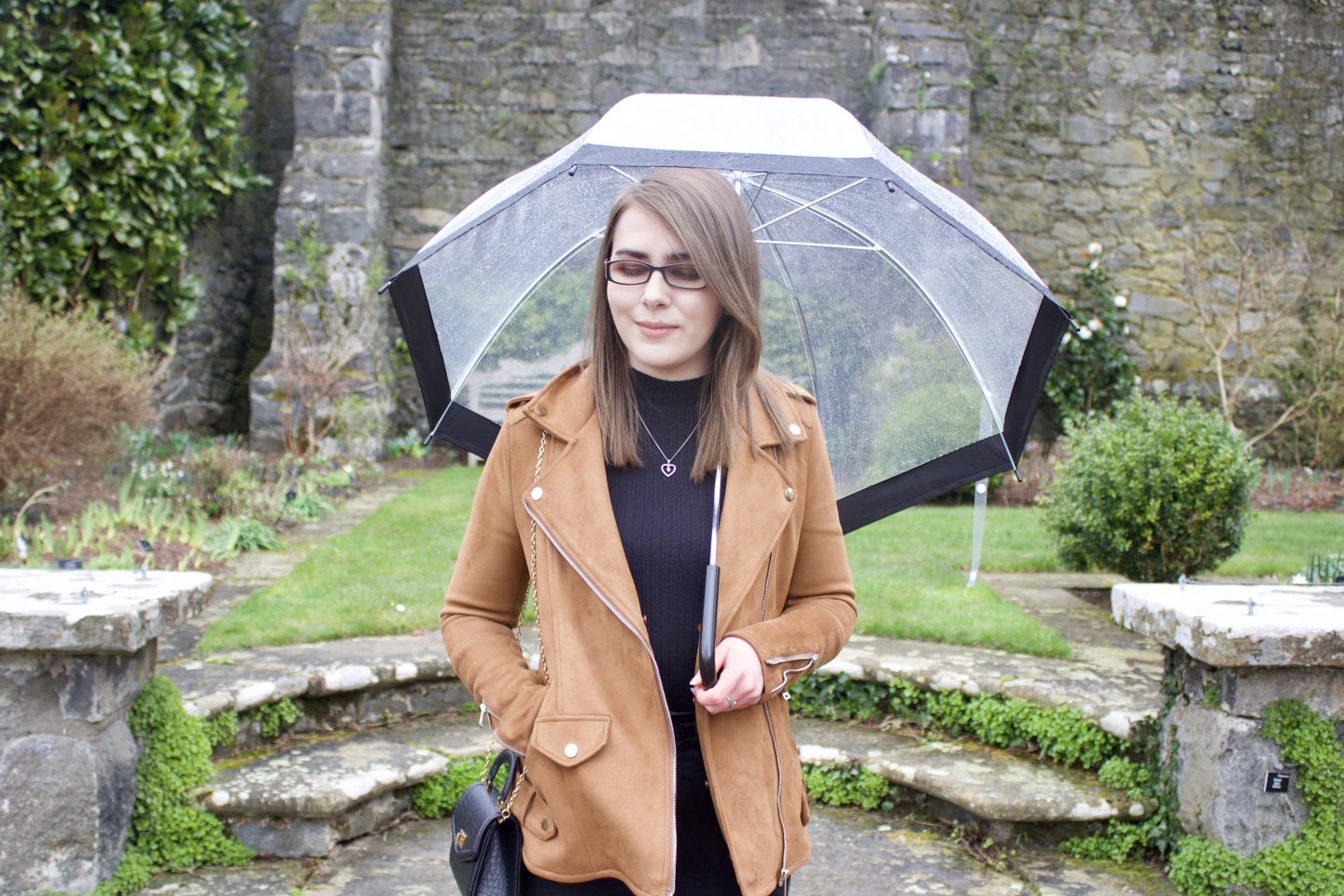 A photo of Elin standing on some stone steps in front of a pretty stone wall with flowers in front of it. She's holding a clear umbrella and looking down
