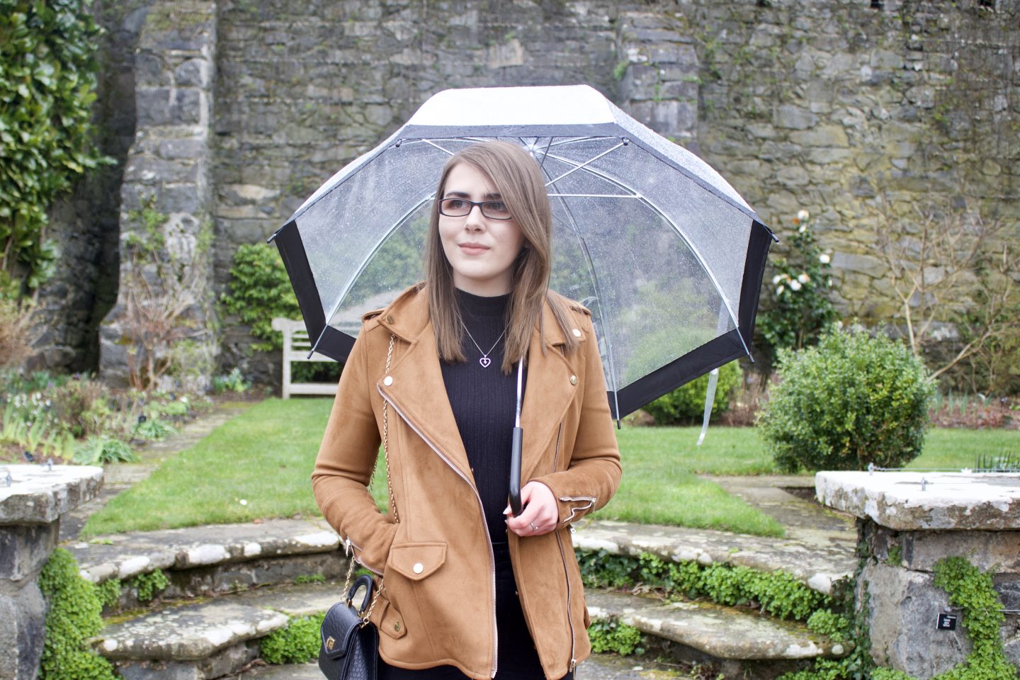 A photo of Elin standing on some stone steps in front of a pretty stone wall which has flowers in front of it. She's looking away from the camera and standing under a clear umbrella