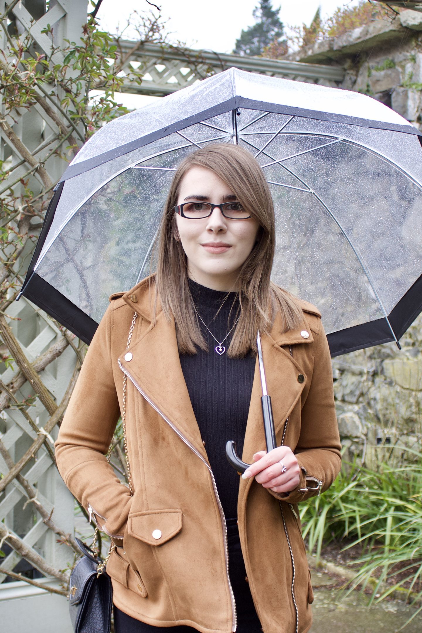 A photo of Elin wearing a black high neck top and black skinny jeans with a brown faux suede jacket. She's also holding a clear umbrella with a black rim