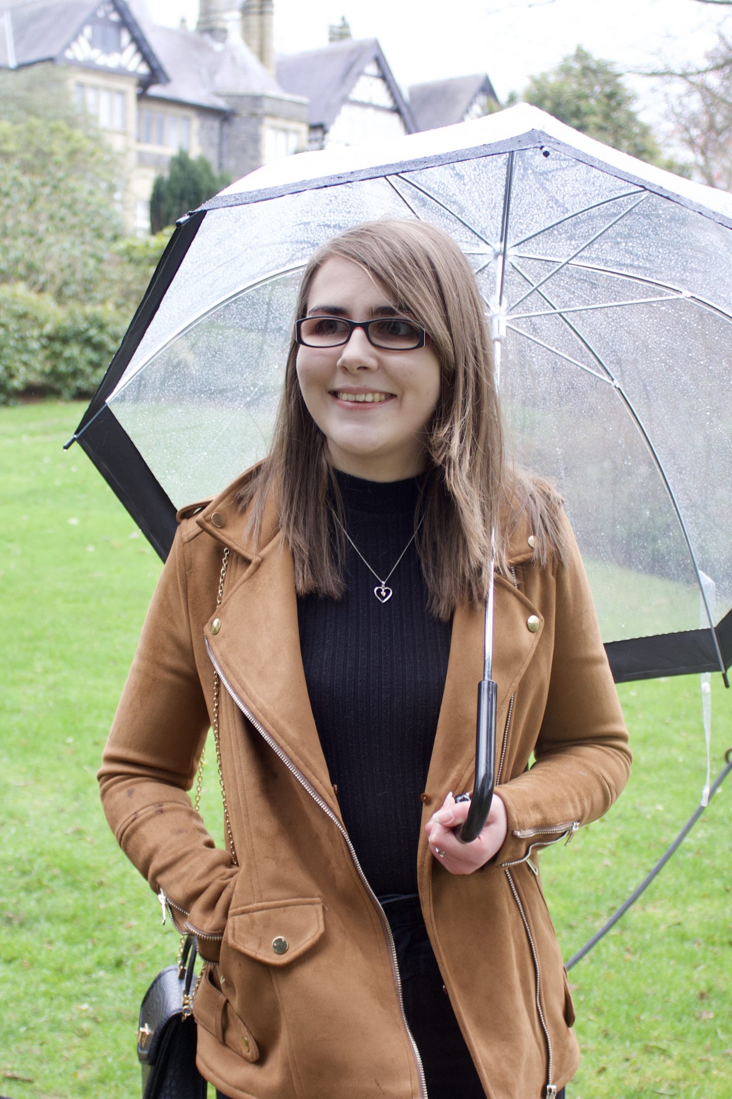 A photo of Elin standing on a lawn with a tudor style house peeking out in the background, she's looking away from the camera and smiling