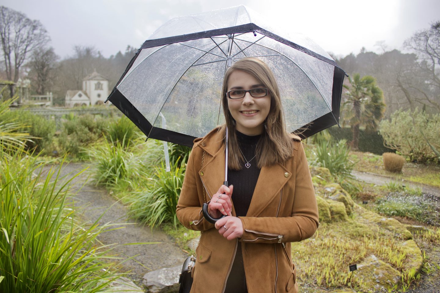 A photo of Elin standing in front of a view of gardens and mountains, she's holding a clear umbrella and smiling at the camera