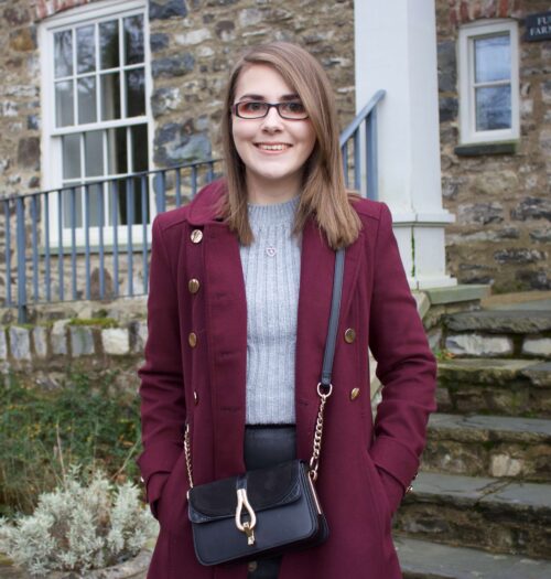 A photo of Elin standing in front of steps that lead up to a pretty stone building. She is wearing a black faux leather skirt, a grey cable knit jumper and a burgundy double breasted longline coat. Her hands are in her pockets and she is smiling at the camera
