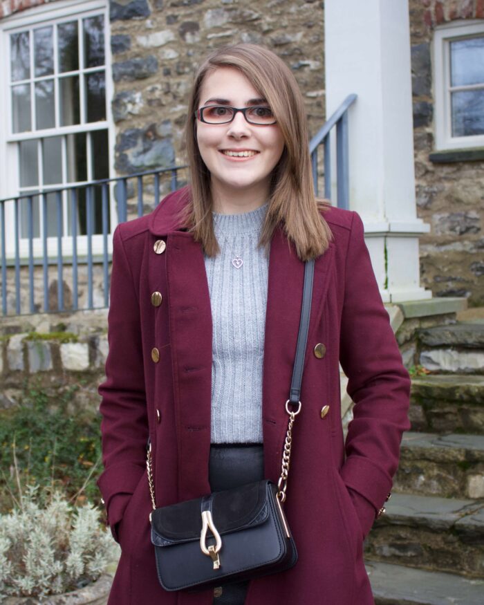 A photo of Elin standing in front of steps that lead up to a pretty stone building. She is wearing a black faux leather skirt, a grey cable knit jumper and a burgundy double breasted longline coat. Her hands are in her pockets and she is smiling at the camera