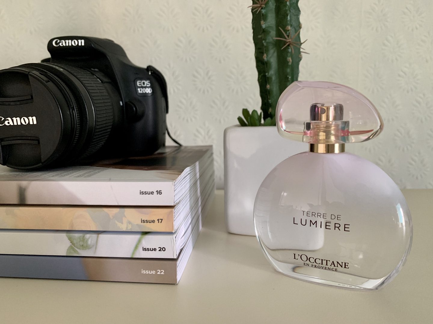 A photo of the Terre de Lumiere perfume by L'occitane in front of a potted fake cactus. A canon camera is placed on top of a stack of Blogosphere Magazines.